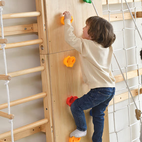 A young child wearing a cream-colored sweater and blue jeans climbs an indoor wooden rock climbing wall with colorful handholds, which is part of the PP AVENLUR Walnut - 9-in-1 Swedish Ladder Wall Gym and Climber that includes a rope net and wooden ladder rungs on the side.