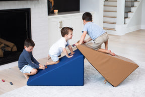 Three young boys play with toy cars on a durable set of Mega Wedge Play Triangles from Foamnasium. One kneels on a blue wedge, another climbs a brown triangle, and the third sits on the floor, moving his car towards the blue wedge. They appear to be in a modern living room with a staircase in the background.